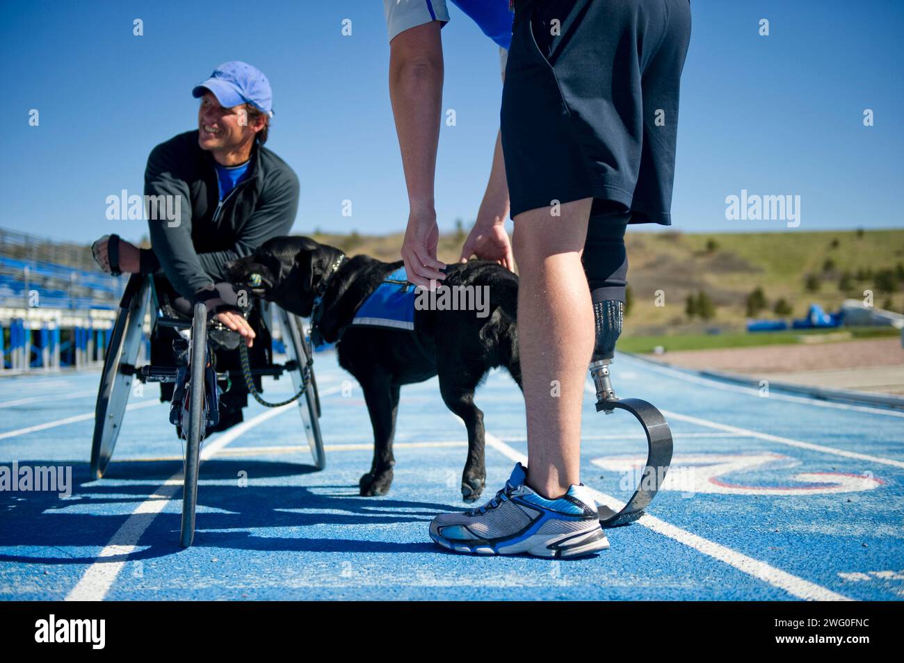 Un athlète avec sa prothèse de course partage un moment avec un athlète en fauteuil roulant et son chien de travail. Banque D'Images