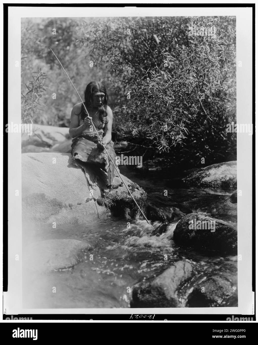 Sur le Merced-Southern Miwok, c1924. Miwok homme tenant la lance, assis sur un rocher dans un ruisseau. Banque D'Images