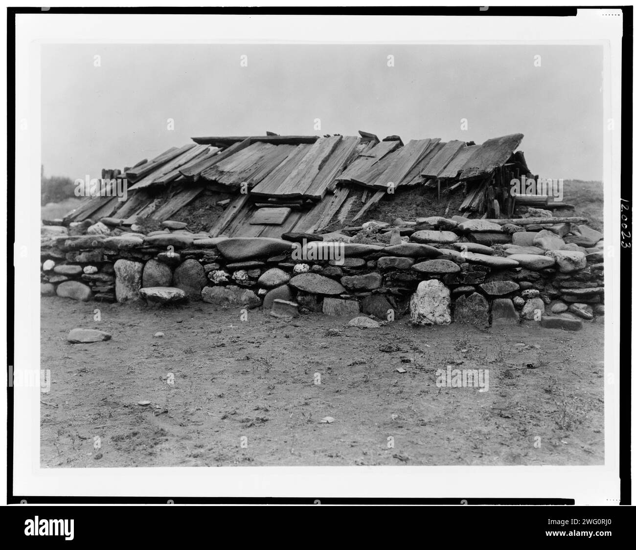 Hupa maison de sueur, 1923. Bâtiment souterrain recouvert d'un toit en planches de bois, entouré d'un mur de gros rochers. Banque D'Images