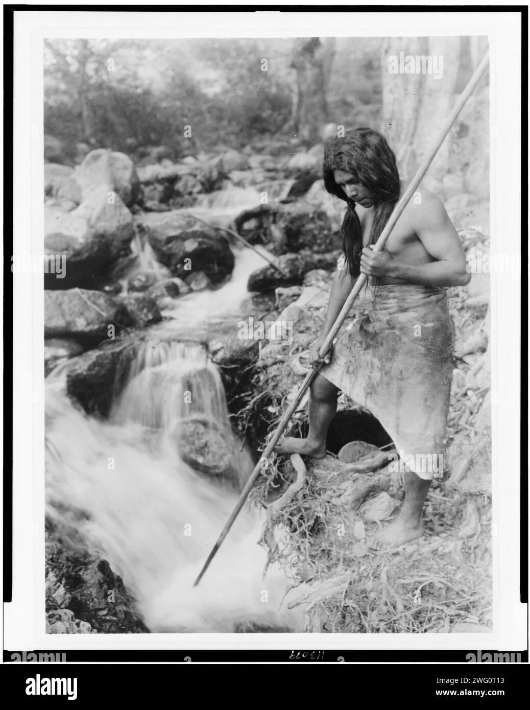 Regarder Salmon-Hupa, c1923. Hupa homme avec lance, debout sur la rive regardant dans le ruisseau. Banque D'Images