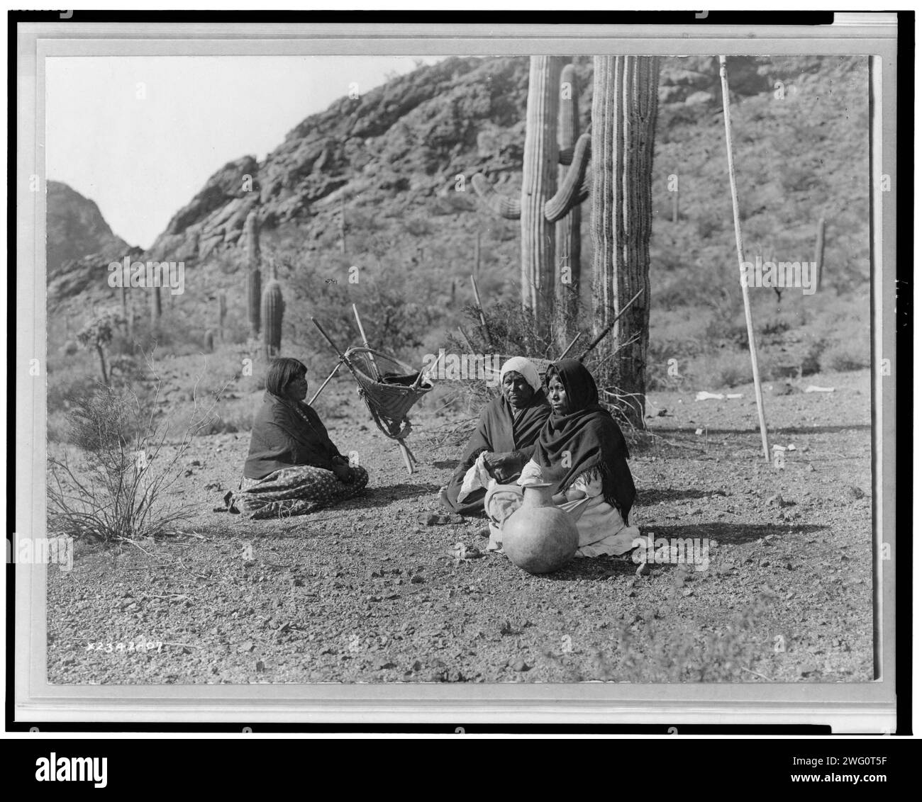Femmes Qahatika se reposant à Harvest Field-Qahatika, c1907. Trois femmes Qahatika assises au sol avec des porte-kiho et du pot à proximité, des cactus saguaro et des montagnes en arrière-plan. Banque D'Images
