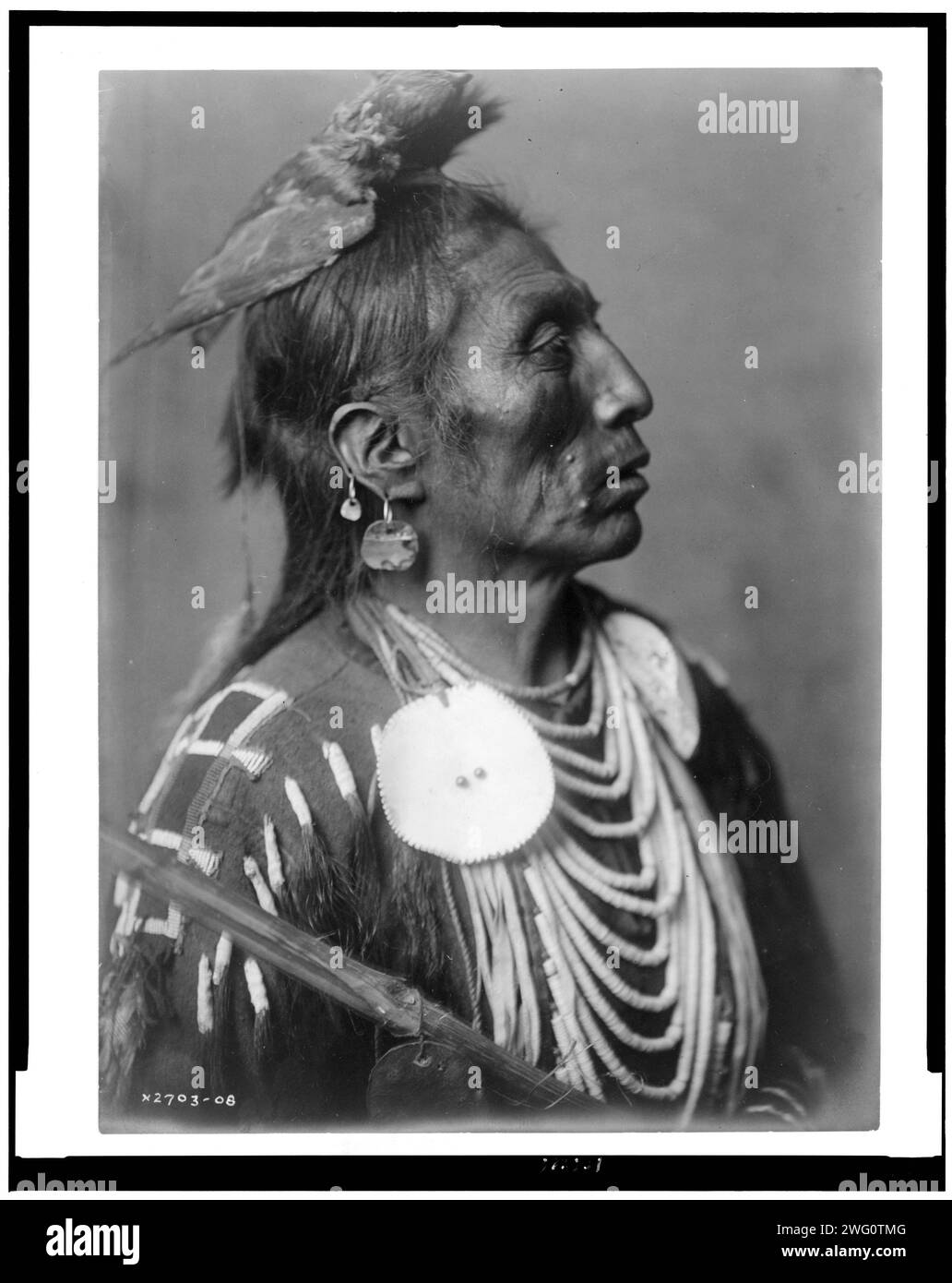 Médecine Crow-Apsaroke, c1908. Medicine Crow, Crow Indian, Montana, portrait tête et épaules, face à droite, coiffe en peau de faucon, deux boucles d'oreilles dans l'oreille droite, ornements de disque, perles de coquille, chemise de cuir chevelu perlé. Banque D'Images