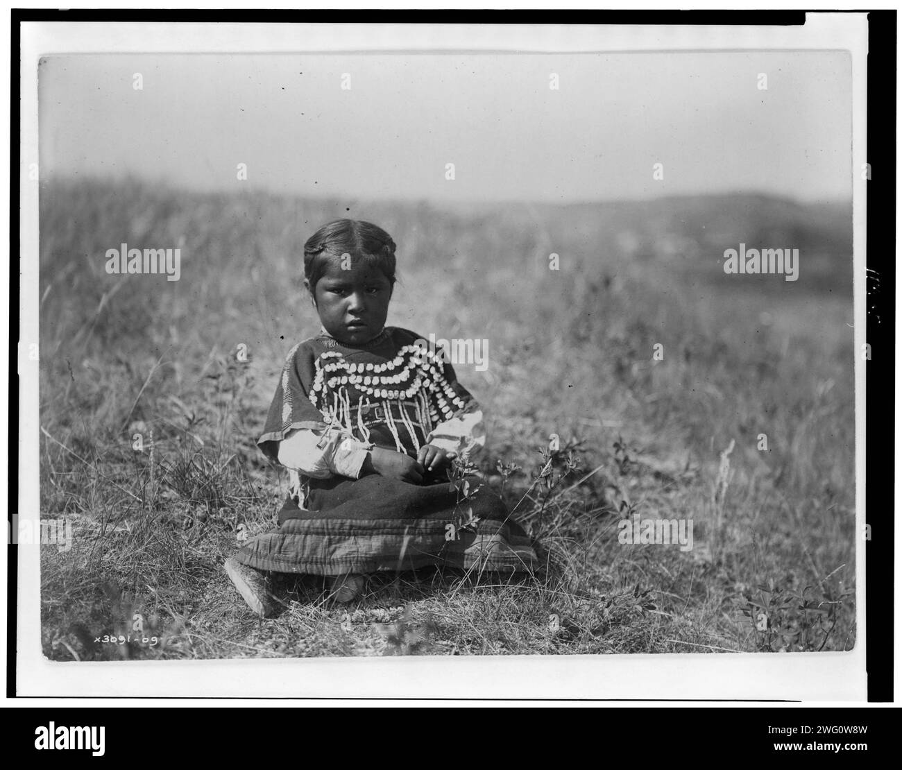 Courir la fille de Owl, c1910. Jeune fille Piegan, portrait en pied, vêtue d'une robe décorée de dents d'élan, assise sur un sol dégagé, face à l'avant. Banque D'Images