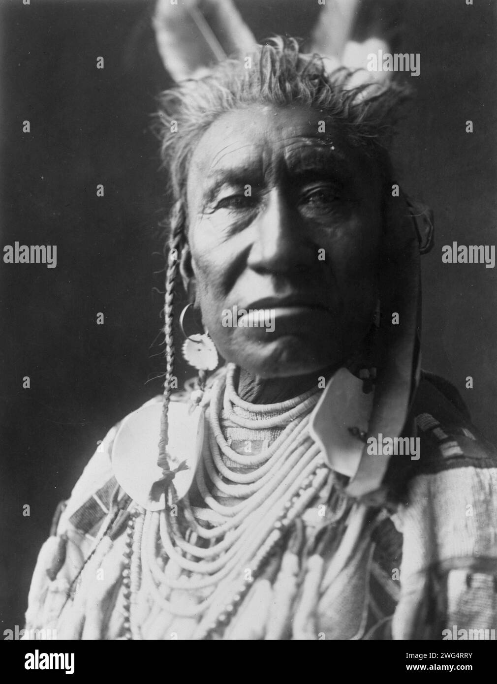 Spectacles de poissons, un Indien Apsaroke, portrait tête et épaules, face à face, pompadour, tresses de temple peintes en blanc, 2 plumes d'aigle, boucle d'oreille disque, perles de coquille avec de grands ornements de disque, chemise en peau de mousseline perlée, c1908. Banque D'Images Spectacles de poissons, un Indien Apsaroke, portrait tête et épaules, face à face, pompadour, tresses de temple peintes en blanc, 2 plumes d'aigle, boucle d'oreille disque, perles de coquille avec de grands ornements de disque, chemise en peau de mousseline perlée, c1908. Banque D'Images