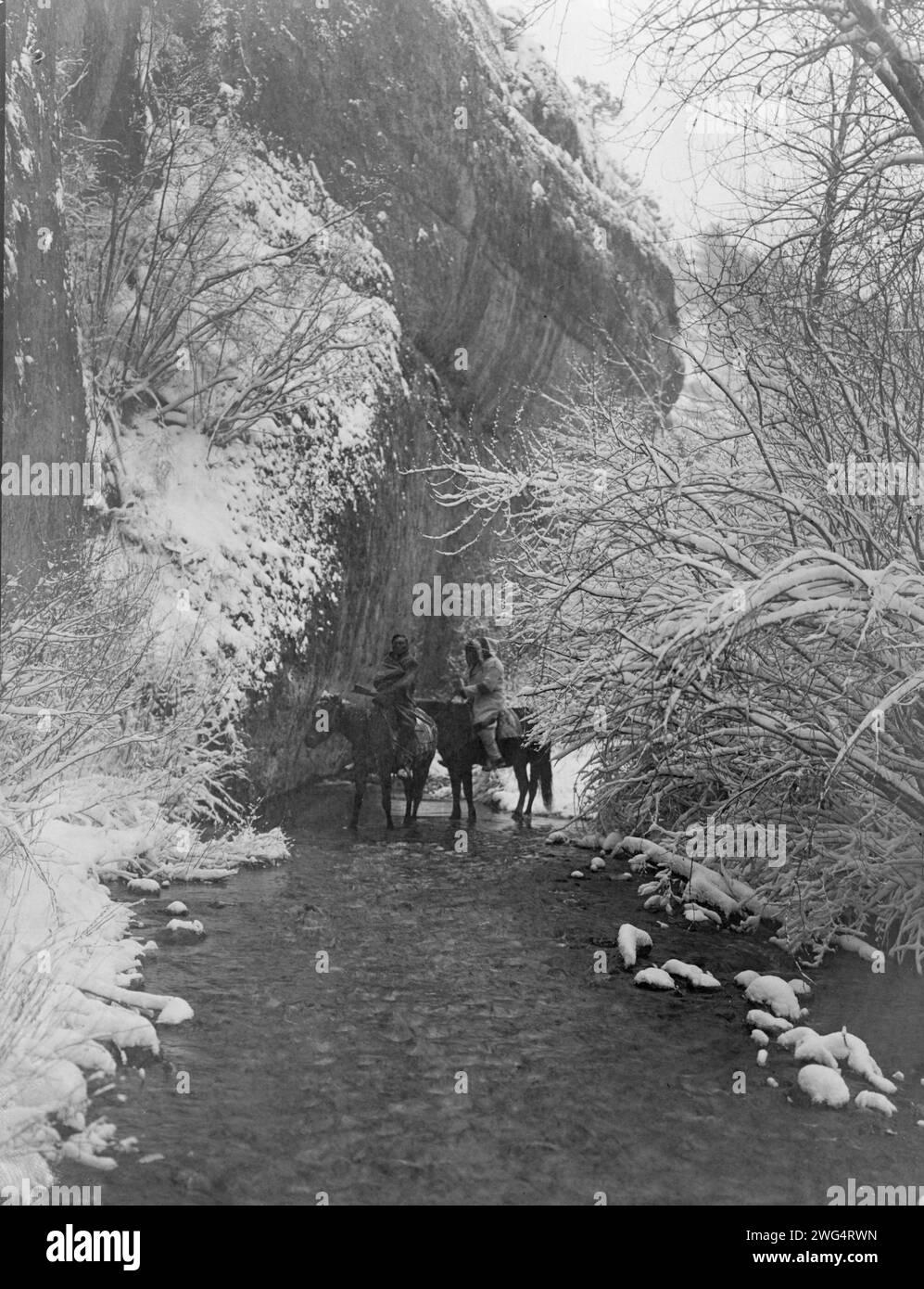 Approche de l'hiver-Apsaroke, c1908. Deux Indiens Crow à cheval dans un ruisseau peu profond flanqué d'arbres enneigés, sous une falaise rocheuse. Banque D'Images