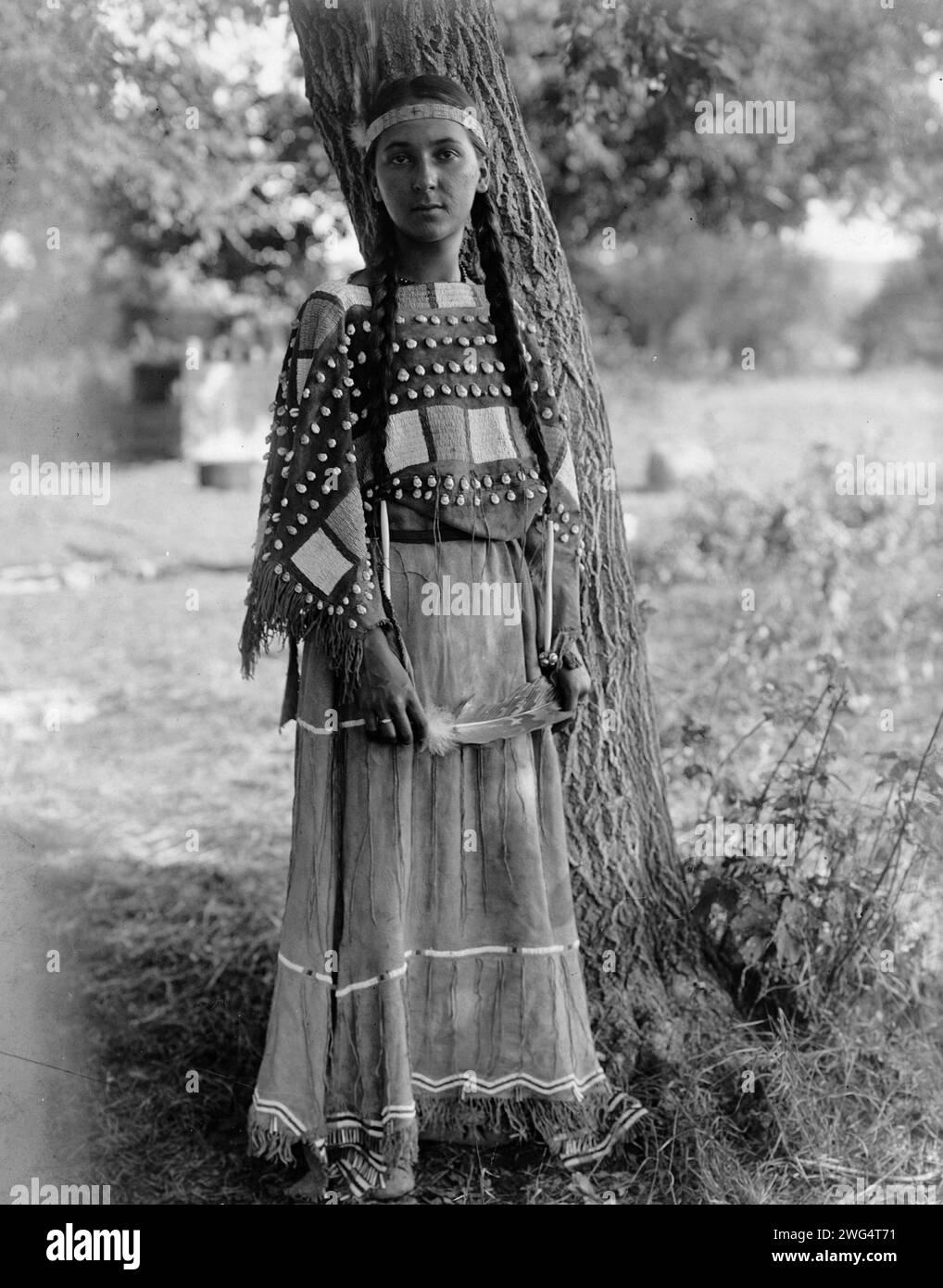 Jeune fille Sioux, c1908. Portrait, pleine longueur, debout près de l'arbre, serre-tête perlé, robe en peau de mousqueton perlée. Banque D'Images