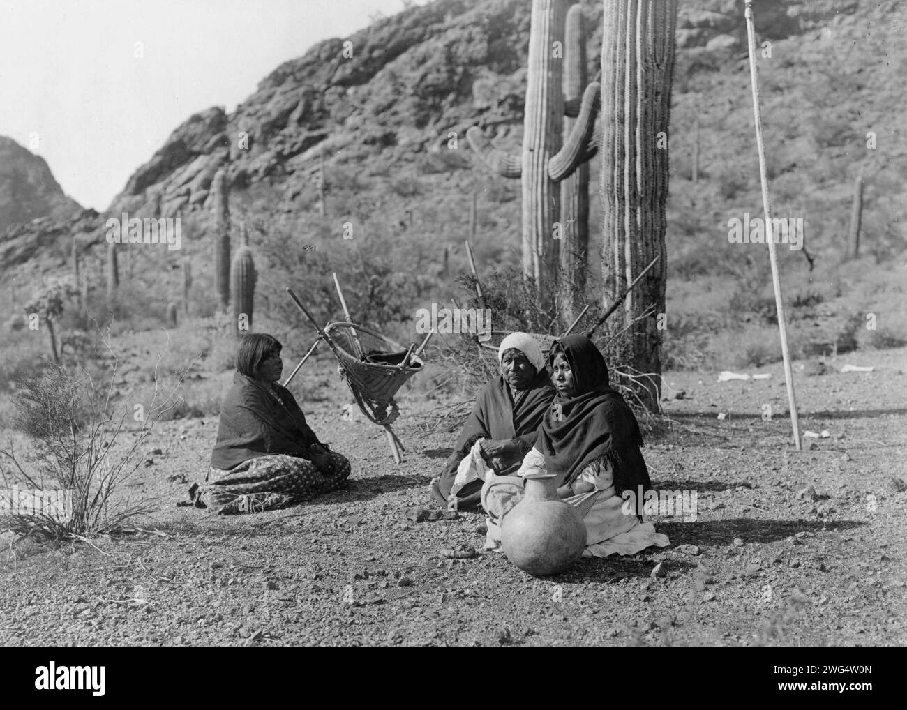 Femmes Qahatika se reposant à Harvest Field-Qahatika, c1907. Trois femmes Qahatika assises au sol avec des porte-kiho et du pot à proximité, des cactus saguaro et des montagnes en arrière-plan. Banque D'Images