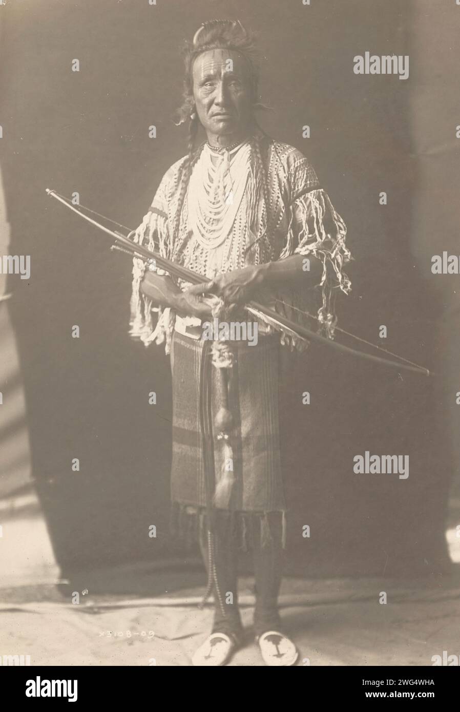 Crow Chief, 1910. La photographie montre le portrait en pied de l'ours grizzli, avec les cheveux tressés, portant des vêtements traidtional et plusieurs colliers, debout, tenant l'arc, face à l'avant. Banque D'Images Crow Chief, 1910. La photographie montre le portrait en pied de l'ours grizzli, avec les cheveux tressés, portant des vêtements traidtional et plusieurs colliers, debout, tenant l'arc, face à l'avant. Banque D'Images