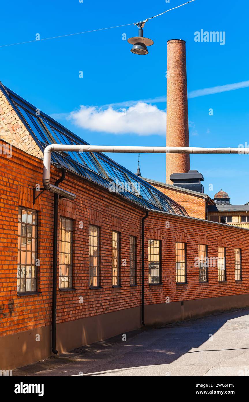 L'image montre un ancien bâtiment d'usine de briques rouges avec une cheminée proéminente sous le ciel bleu clair, indiquant une journée de beau temps. Banque D'Images