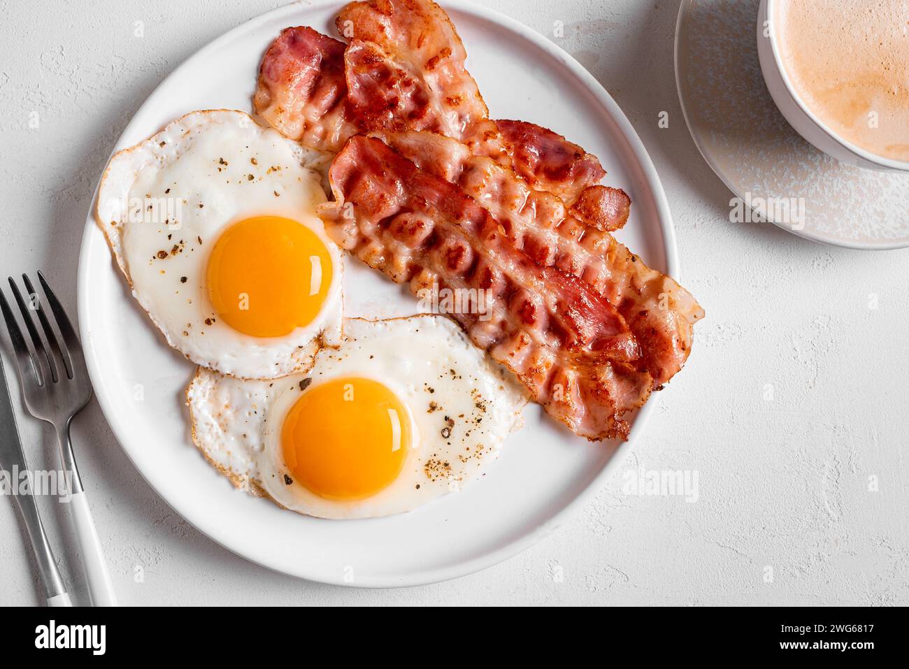 Petit déjeuner avec œufs au plat, bacon et café sur table blanche, vue de dessus. Œufs de côté ensoleillés frits avec bacon sur plaque céto et café cappuccino du matin. Banque D'Images