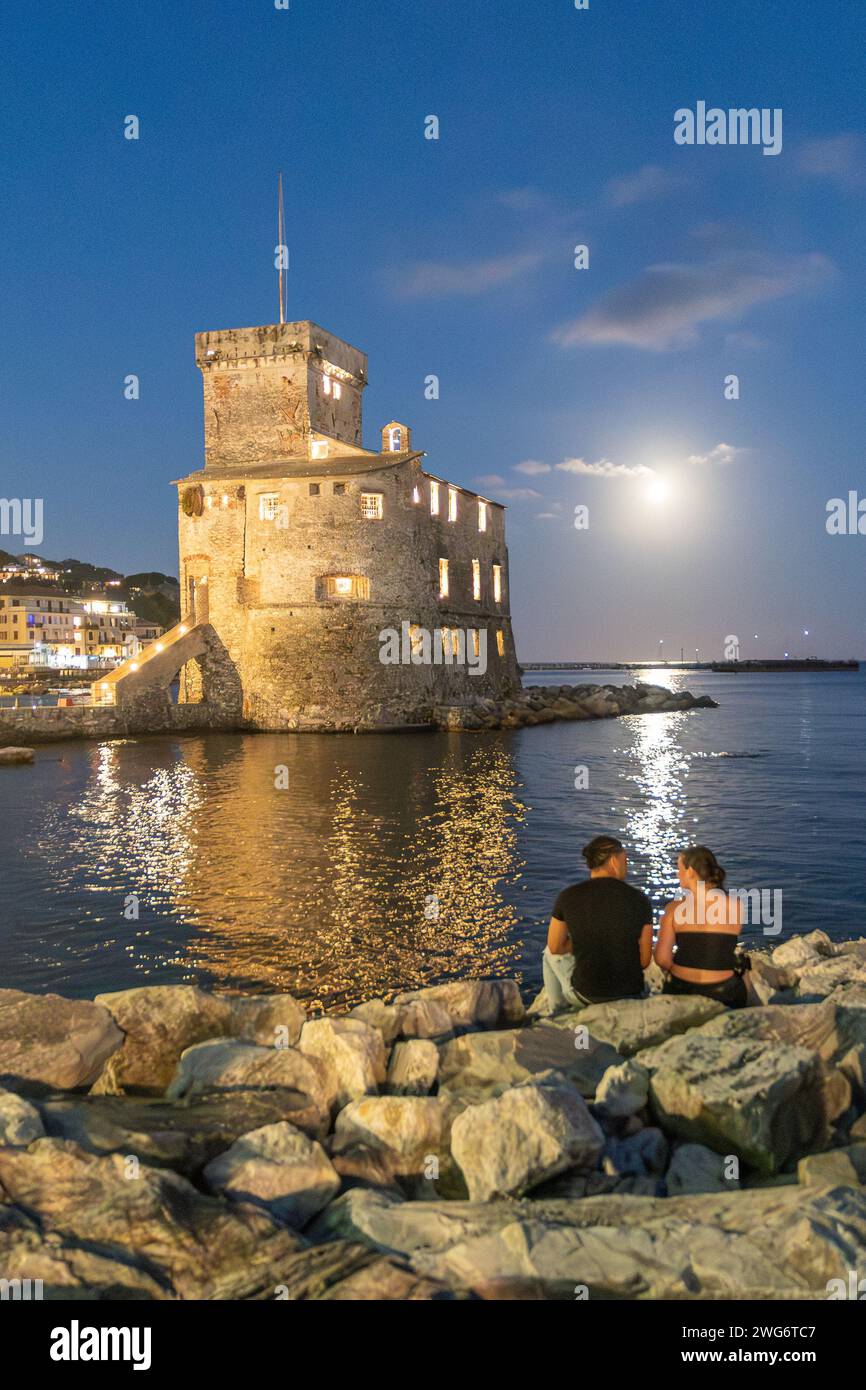 Un jeune couple assis sur la plage de roche en face du château de bord de mer illuminé dans une nuit de pleine lune en été, Rapallo, Gênes, Ligurie, Italie Banque D'Images