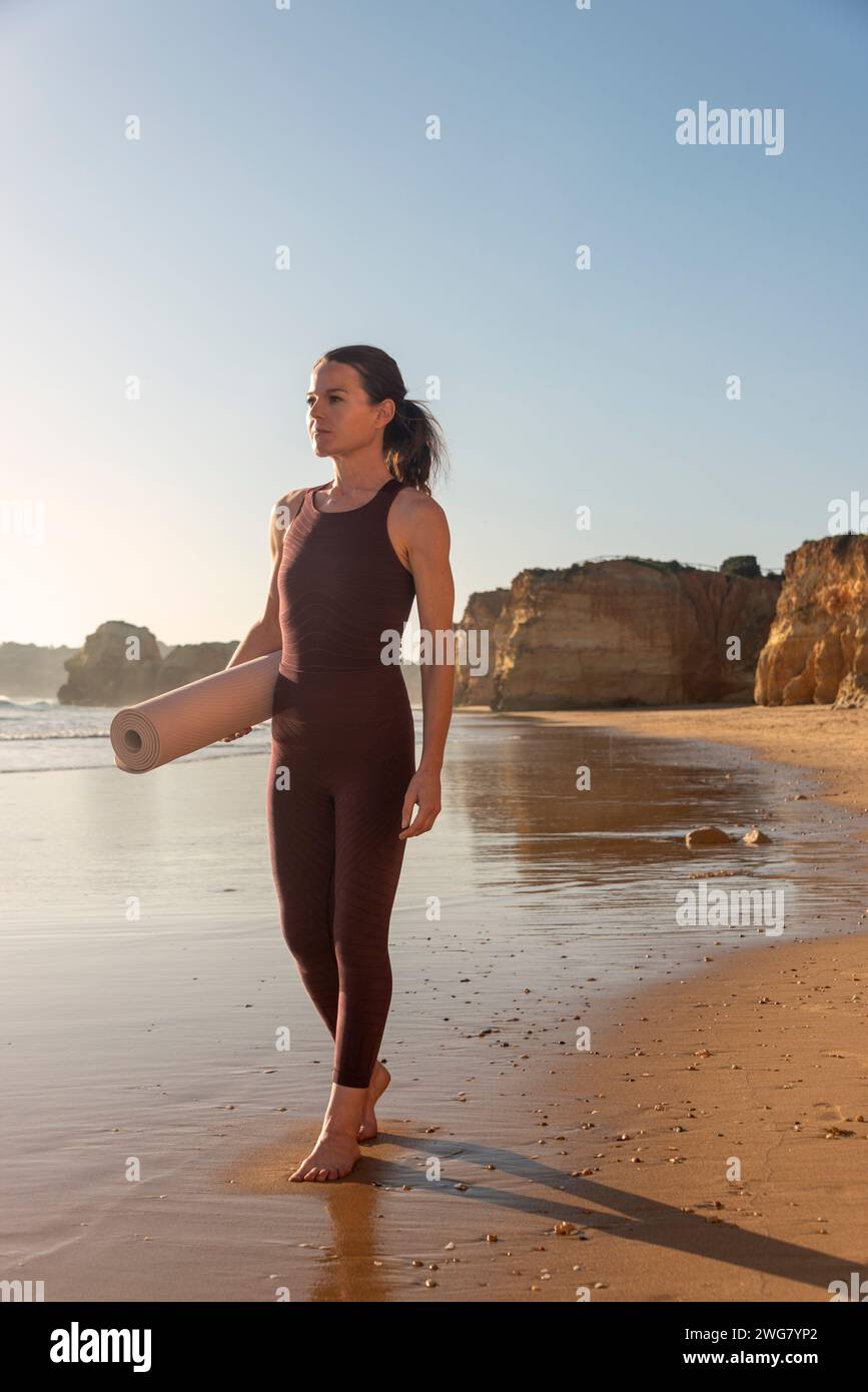 Femme sportive marchant le long d'une plage tenant un tapis d'exercice Banque D'Images