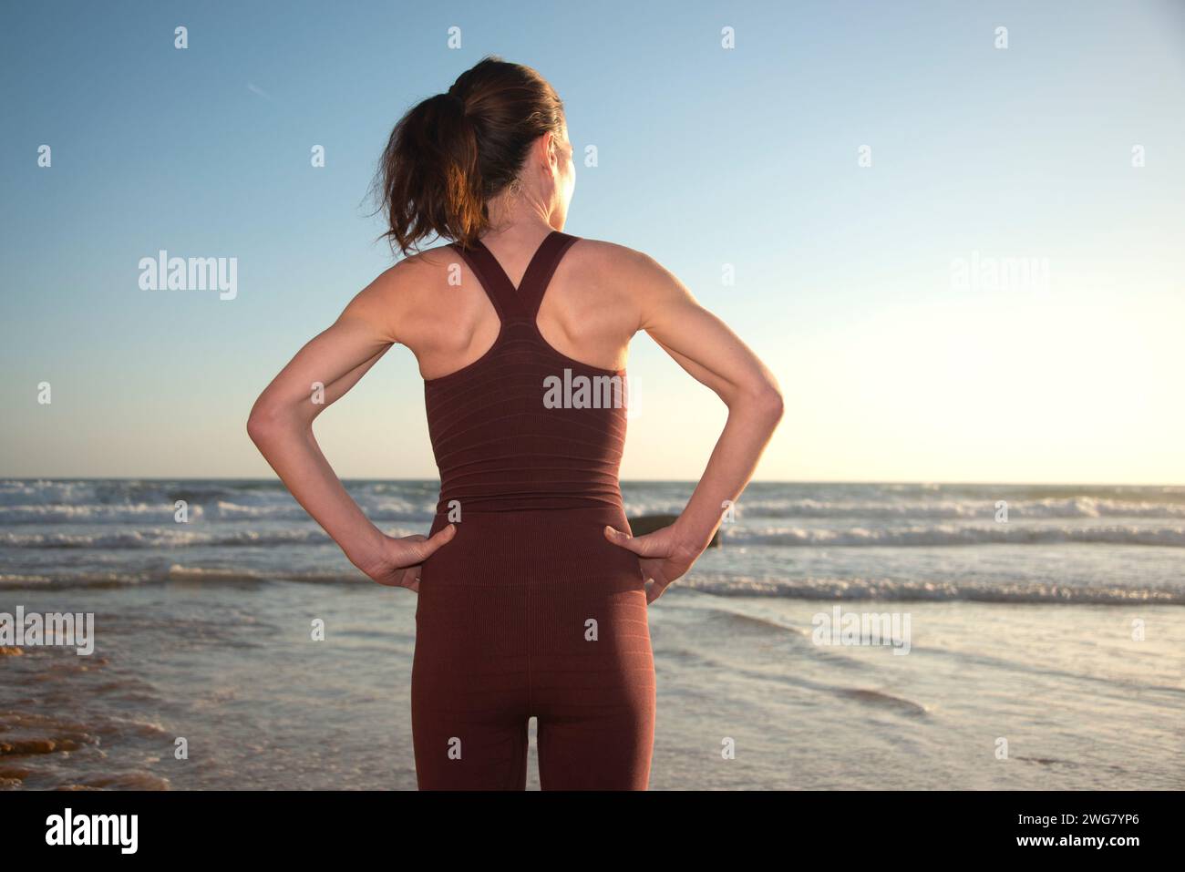 Vue arrière d'une femme sportive face à l'océan avec les mains sur les hanches, nageuse. Banque D'Images