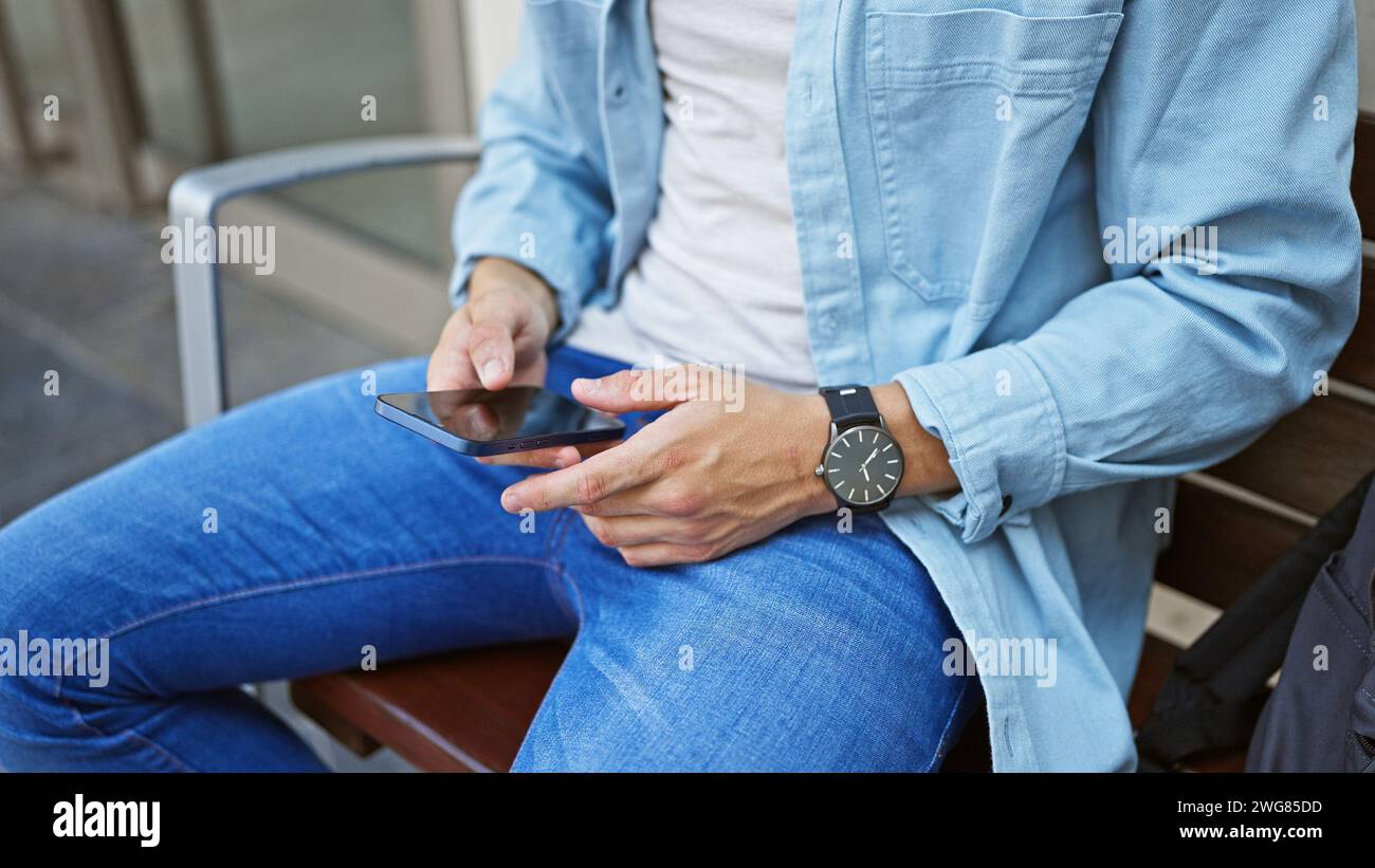 Un homme élégant en tenue décontractée utilise un smartphone assis sur un banc urbain, représentant la connectivité moderne. Banque D'Images