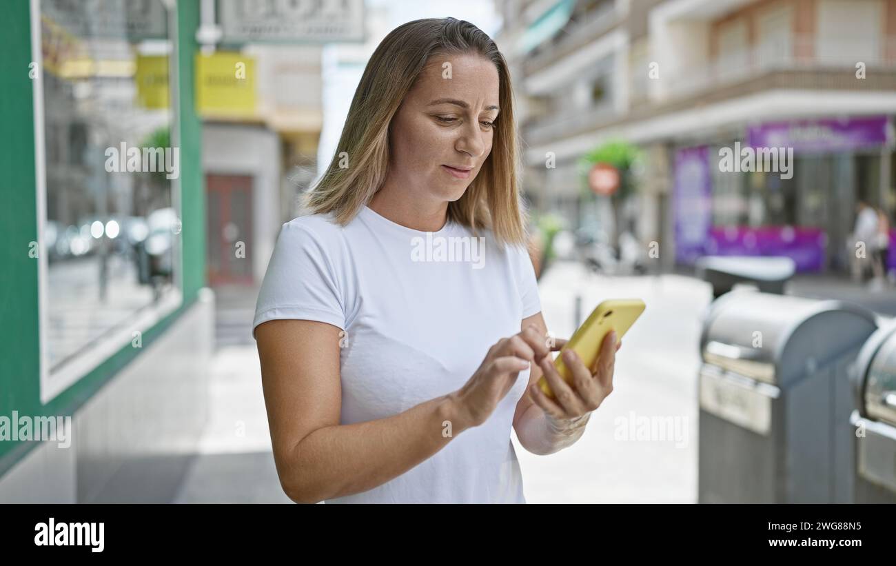 Élégant belle jeune femme blonde sérieusement concentrée sur SMS message en ligne sur son smartphone, debout sur la rue ensoleillée de la ville à l'extérieur, livin Banque D'Images