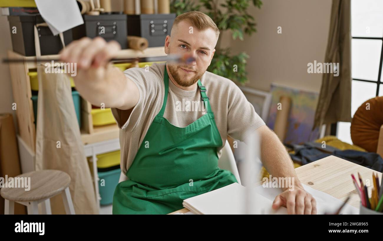 Un jeune homme barbu dans un tablier vert mesure avec un pinceau dans un studio d'art. Banque D'Images