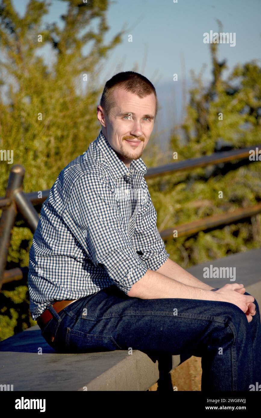 Visiteur assis sur un banc de béton au sommet de Pilot Butte dans l'Oregon. Il porte une chemise bleu marine à carreaux et un Jean bleu. Il sourit. Banque D'Images