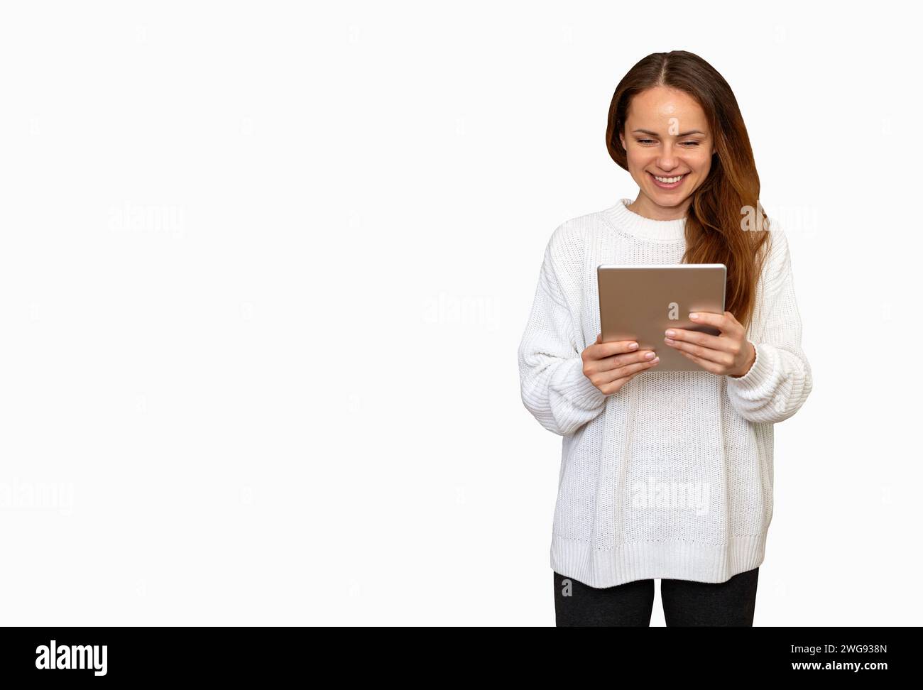 Isolée jeune femme brune avec les cheveux longs portant un pull blanc à l'aide d'une tablette numérique et souriant. Banque D'Images