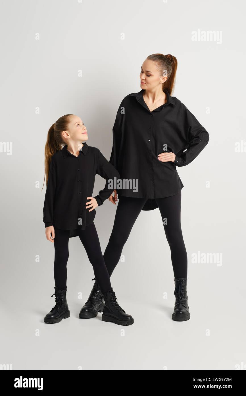 Une famille moderne en tenue noire totale. La mère et sa fille portent des bottes noires assorties, des chemises et des pantalons serrés qui se regardent l'une l'autre Banque D'Images