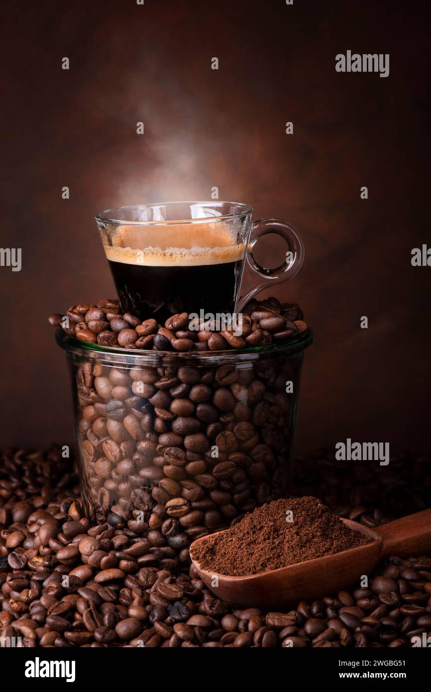 un bocal en verre rempli de grains de café torréfiés, sur une tasse d'espresso à la vapeur. Banque D'Images