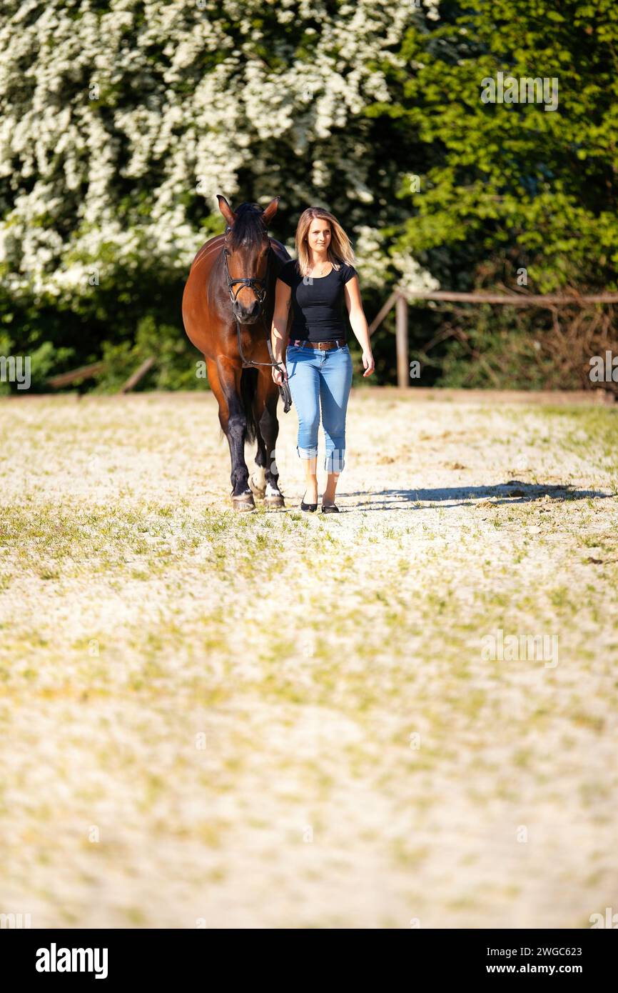 Une jeune femme se tient debout avec son cheval sur une arène d'équitation ensoleillée. En arrière-plan, arbres en fleurs et nature verte, modèle isolé devant le flou, bro Banque D'Images