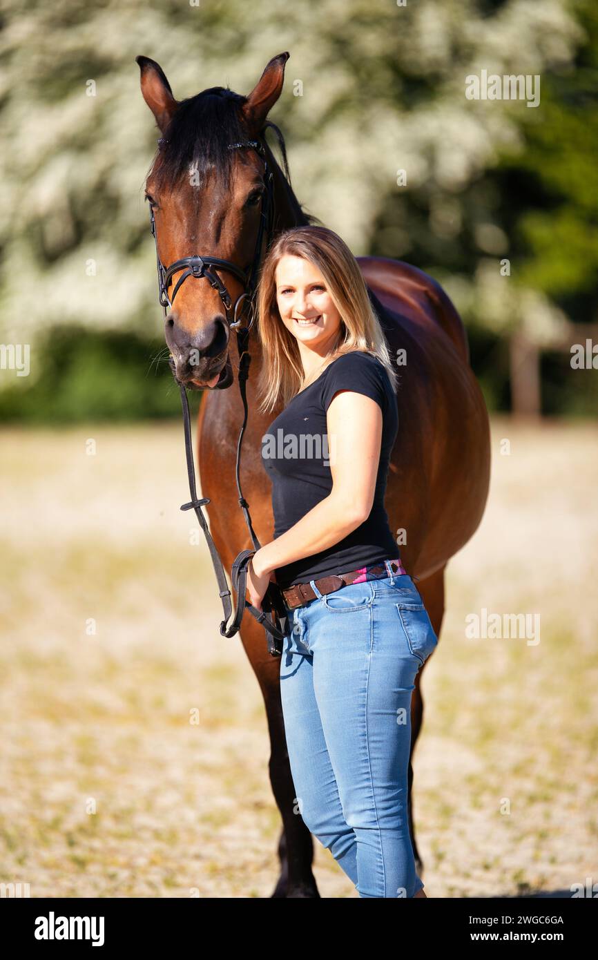 Une jeune femme se tient debout avec son cheval sur une arène d'équitation ensoleillée. En arrière-plan, arbres en fleurs et nature verte, modèle isolé devant le flou, bro Banque D'Images