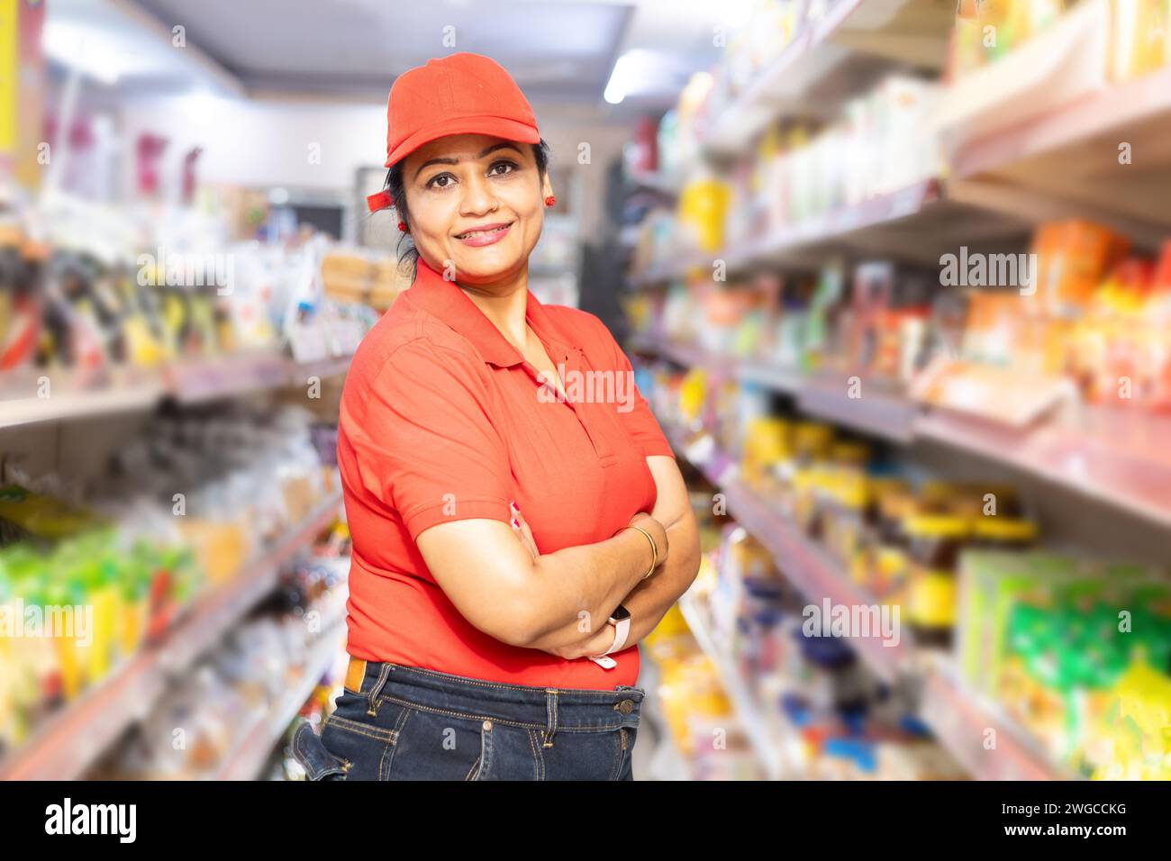 Heureux jeune indien personnel de magasin d'épicerie portant T-shirt rouge et casquette debout avec des bras croisés dans le supermarché. Concept d'autonomisation des femmes Banque D'Images