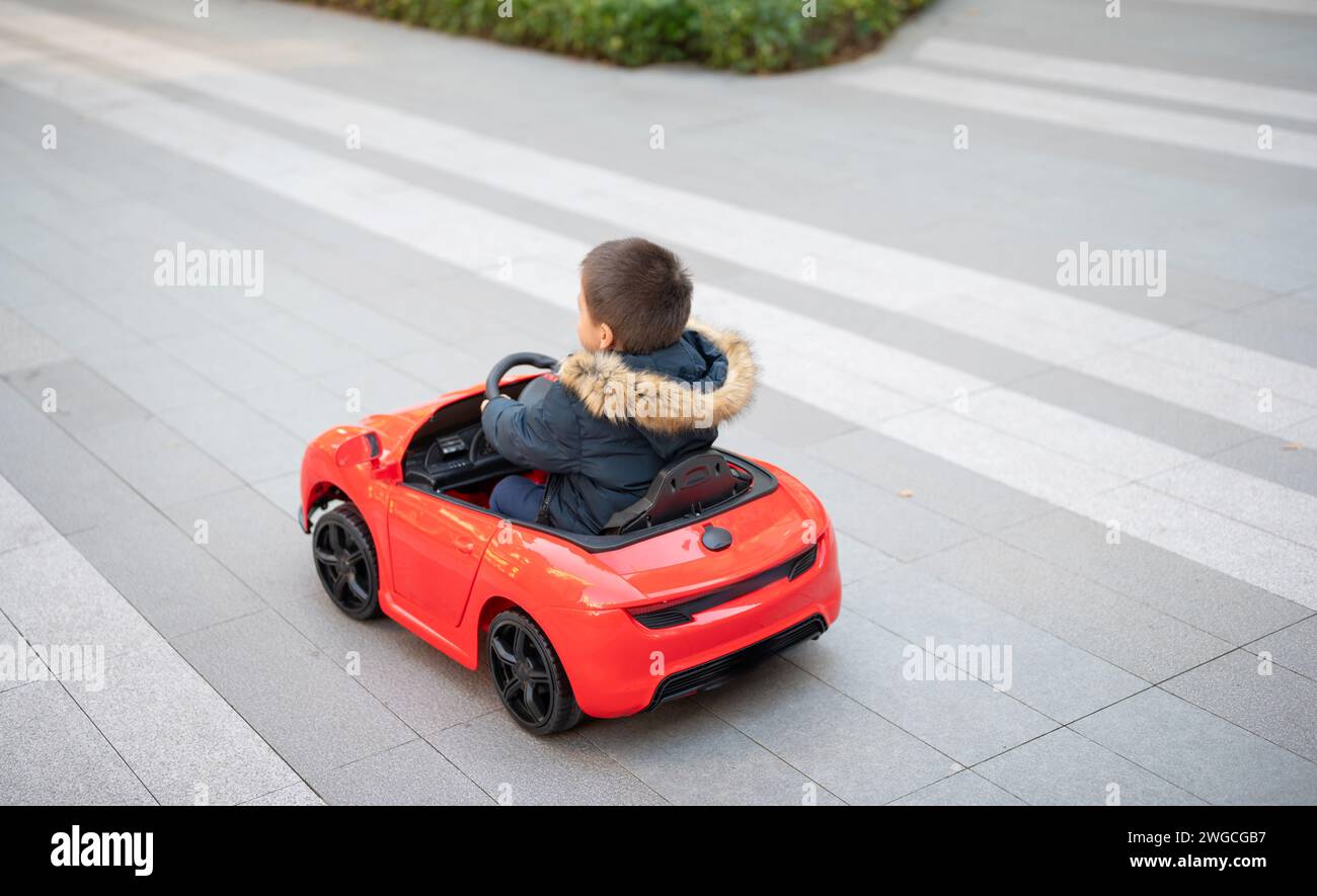 Un jeune enfant multiracial navigue joyeusement la route du quartier dans une voiture de sport rouge jouet, enfilant un grand sourire alors qu'ils prennent le contrôle de leur jouet ca Banque D'Images
