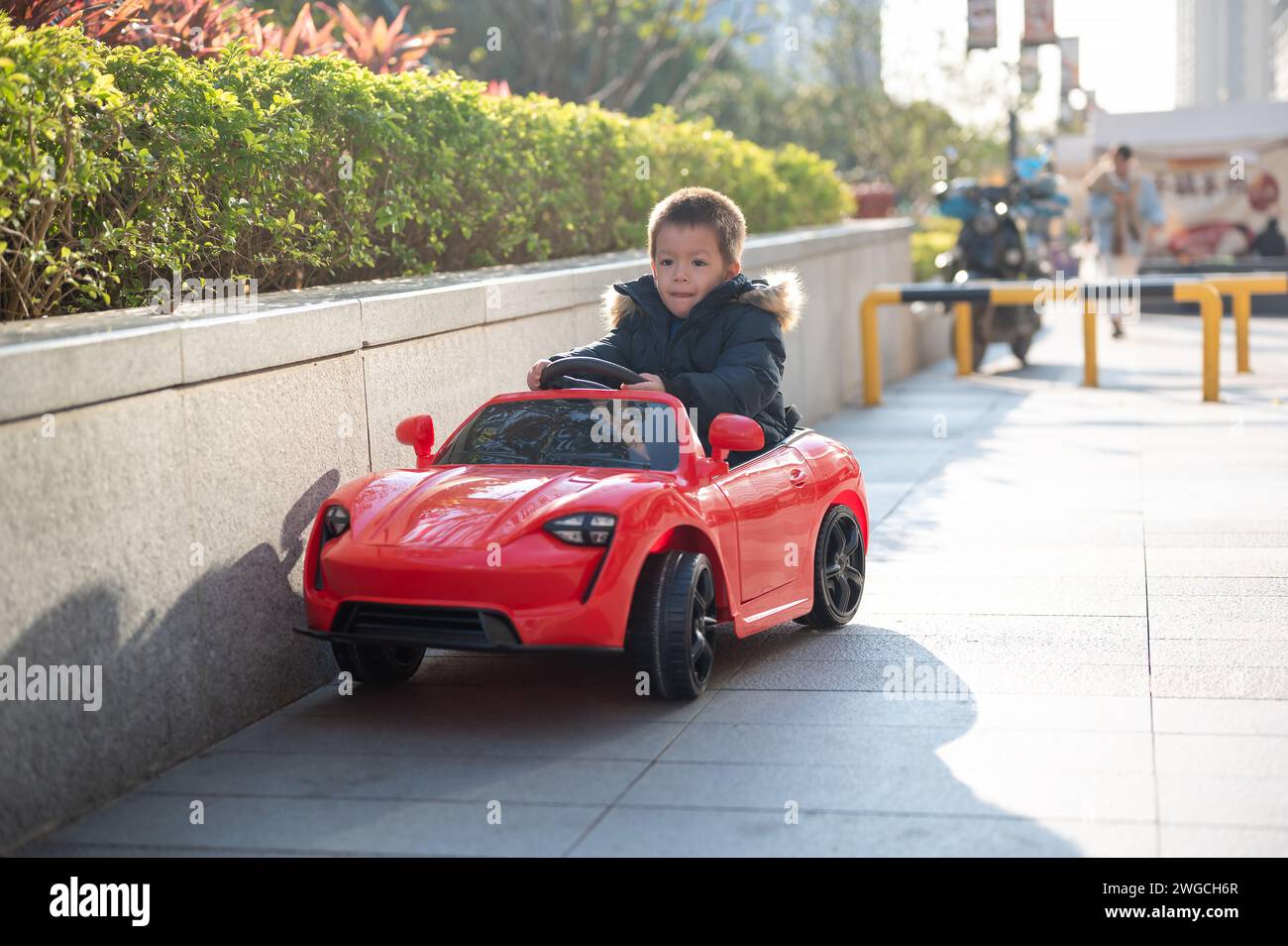 Un jeune enfant multiracial navigue joyeusement la route du quartier dans une voiture de sport rouge jouet, enfilant un grand sourire alors qu'ils prennent le contrôle de leur jouet ca Banque D'Images