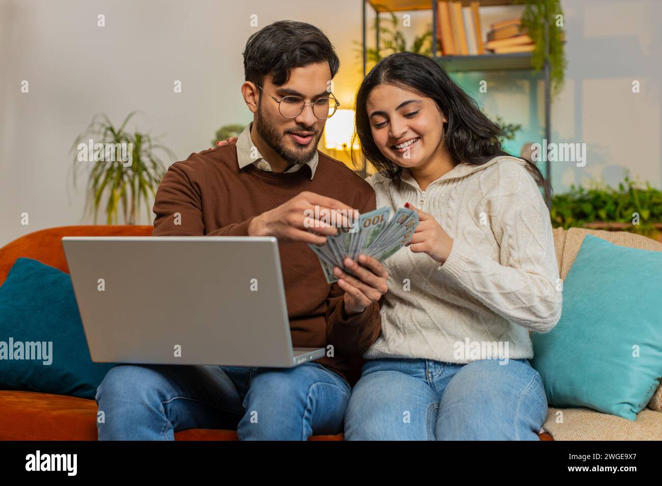 Heureux jeune couple indien comptant de l'argent et des billets de réservation pour des vacances se trouve sur le canapé dans le salon à la maison. Famille diversifiée détenant des billets de banque planification de budget d'investissement en ligne Home Finance Banque D'Images