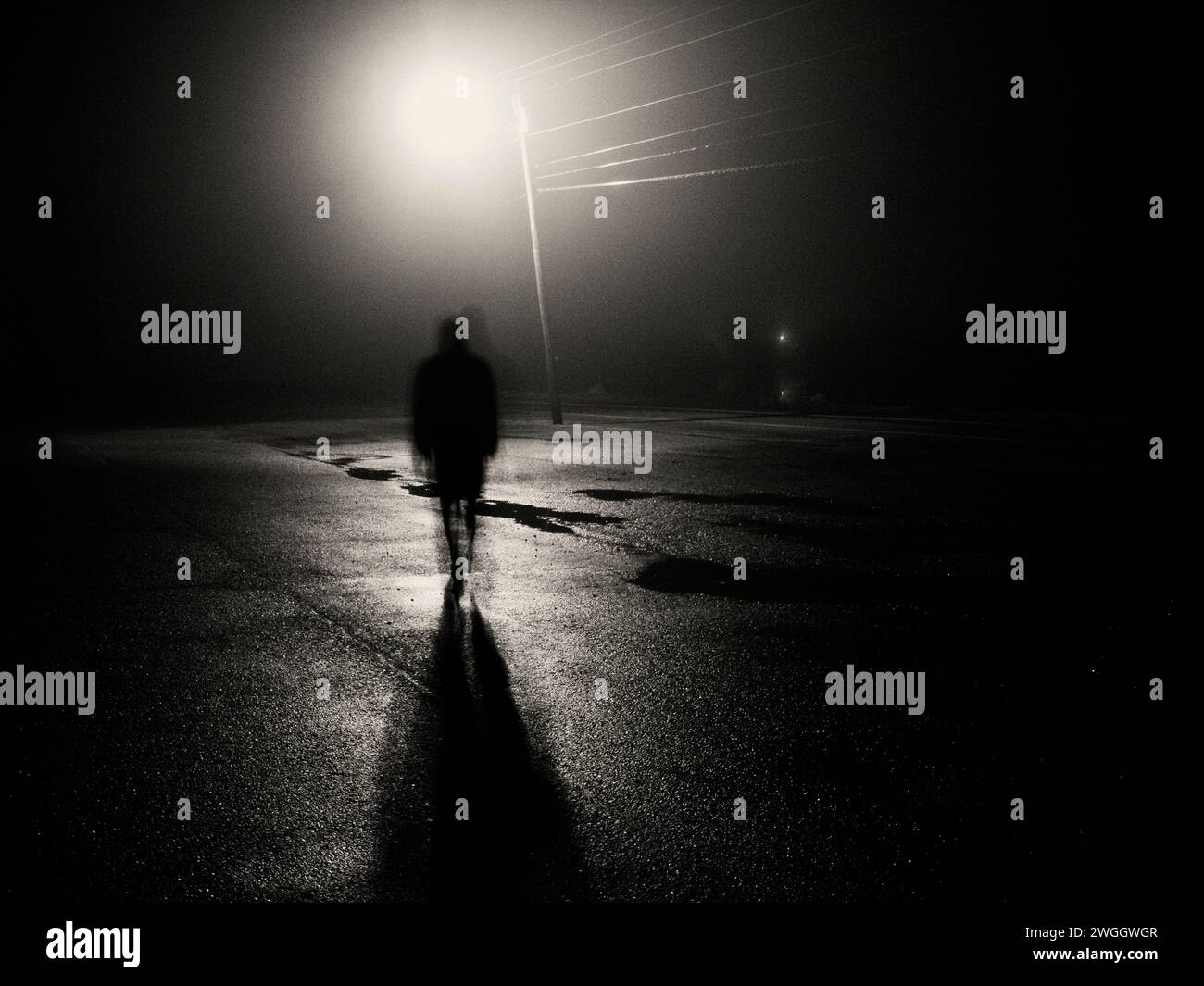 Un personnage ombragé marche à travers un parking trempé de pluie sous un lampadaire la nuit. Banque D'Images