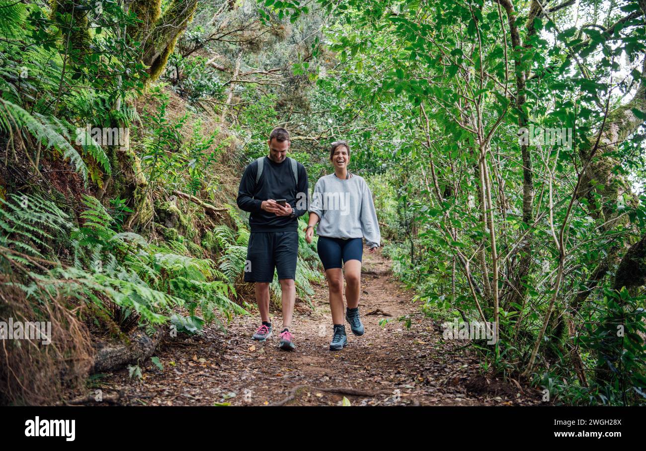Un jeune couple sportif marche sur un sentier à travers la forêt en regardant leur téléphone Banque D'Images