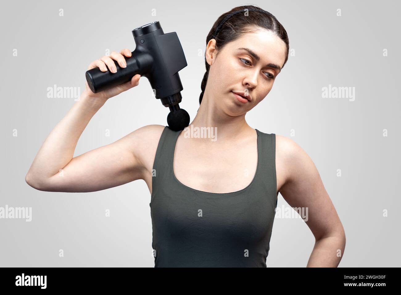Portrait de jeune femme caucasienne se donne massage sur le bras supérieur avec masseur-pistolet à percussion avec buse à bille. Fond de studio gris. Thérapie fo Banque D'Images
