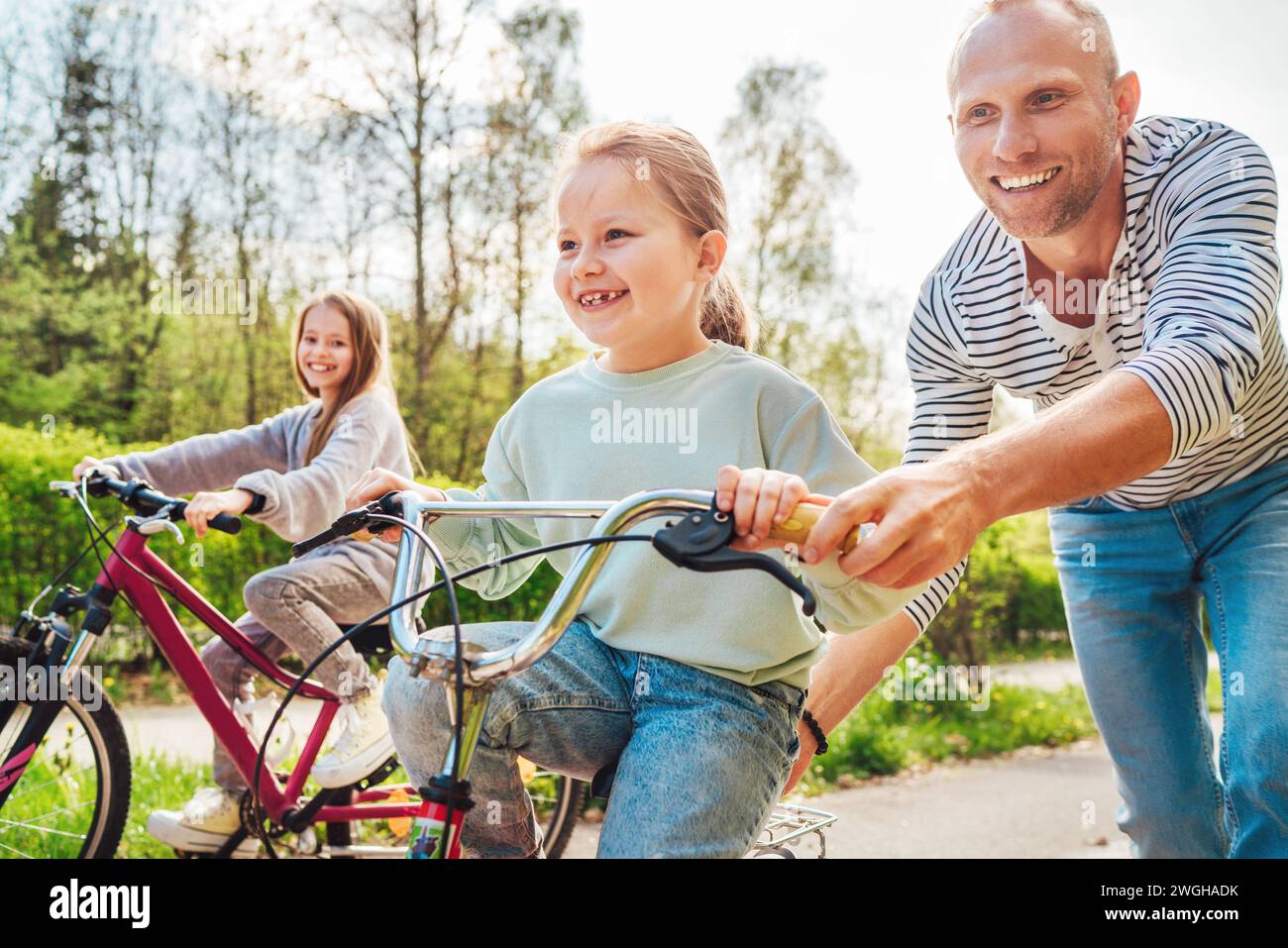 Père souriant avec deux filles pendant la promenade en plein air. Il enseigne à une jeune fille de faire du vélo. Ils apprécient la convivialité dans le parc de la ville d'été. Ha Banque D'Images