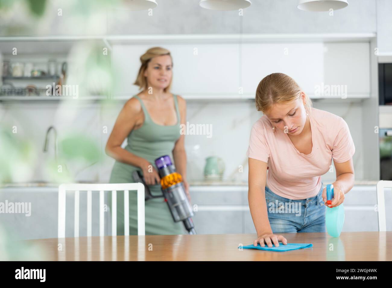 Adolescente aider la mère et laver la table à manger dans la cuisine, nettoyage flou maman à l'aide d'un aspirateur Banque D'Images