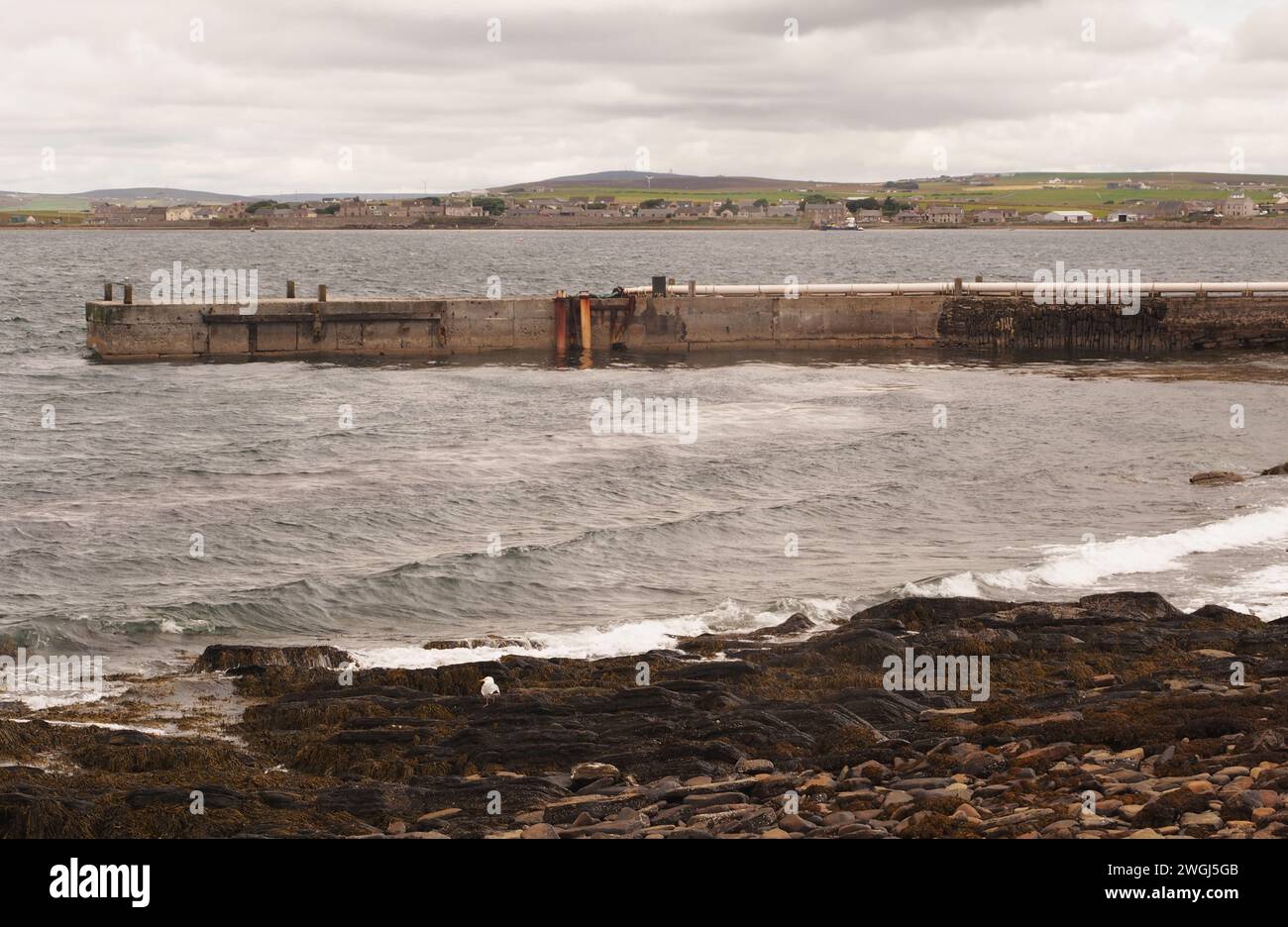 Vue depuis Churchill Barrier 2, sur un petit quai et sur St Mary's Bay jusqu'à St Mary's, Orcades, avec des vagues et la plage au premier plan. Banque D'Images