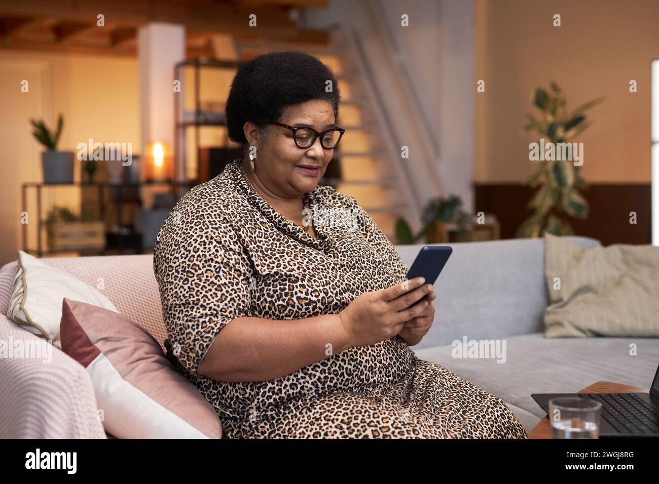 Portrait de femme senior noire moderne portant la robe imprimée guépard à l'aide d'un smartphone assis sur un canapé dans l'espace de copie intérieur de la maison Banque D'Images