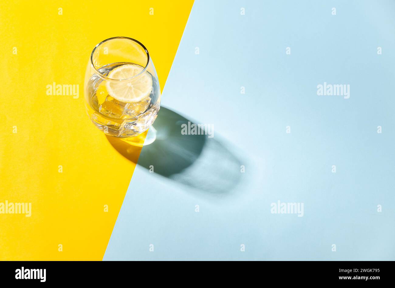Un verre d'eau avec une tranche de citron et des glaçons, sur un fond jaune et bleu avec espace de copie. Banque D'Images