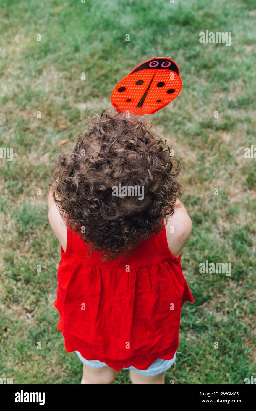 enfant dans une chemise rouge avec des cheveux bruns bouclés tenant une tapette de mouche coccinelle rouge sur l'herbe verte Banque D'Images