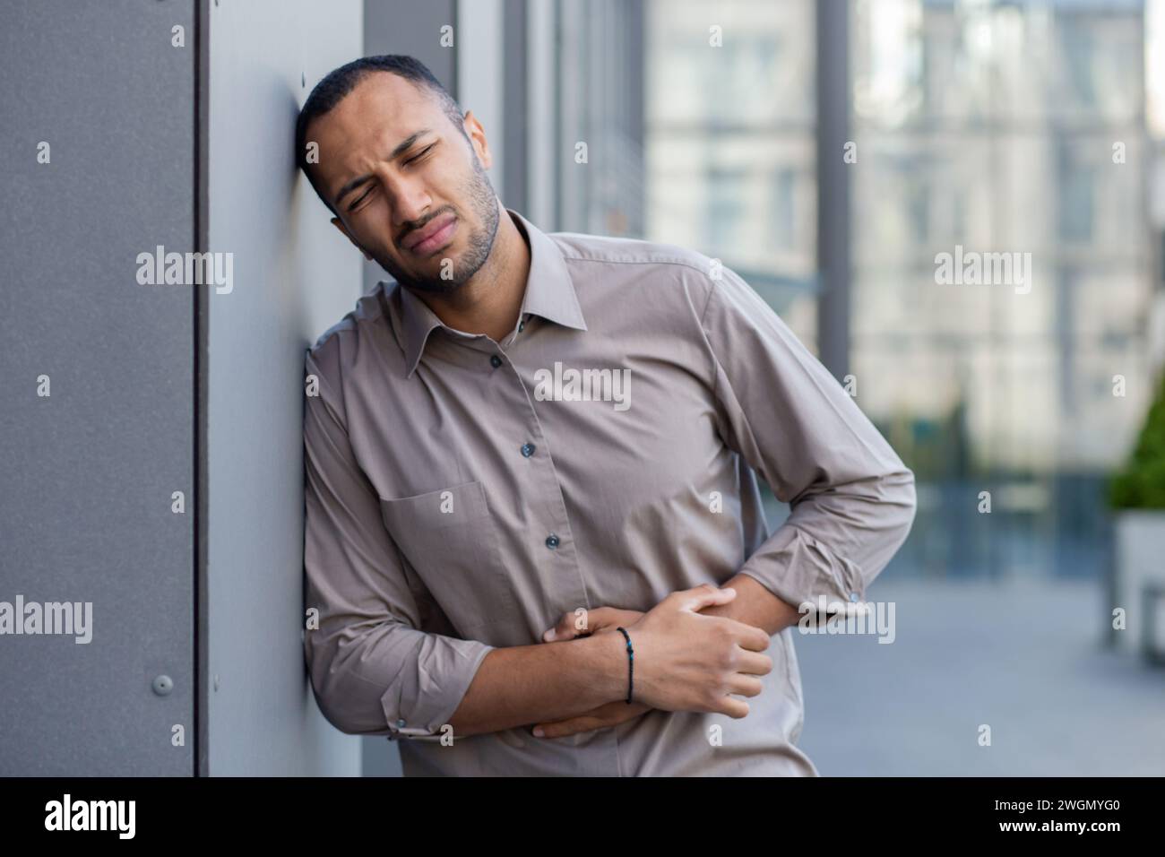 Mâle hispanique malade en vêtements de forme ayant mal au ventre et fermant les yeux dans la douleur tout en se tenant debout en ville. Homme d'affaires épuisé tenant les mains sur le ventre et souffrant de spasmes dus à une intoxication alimentaire. Banque D'Images