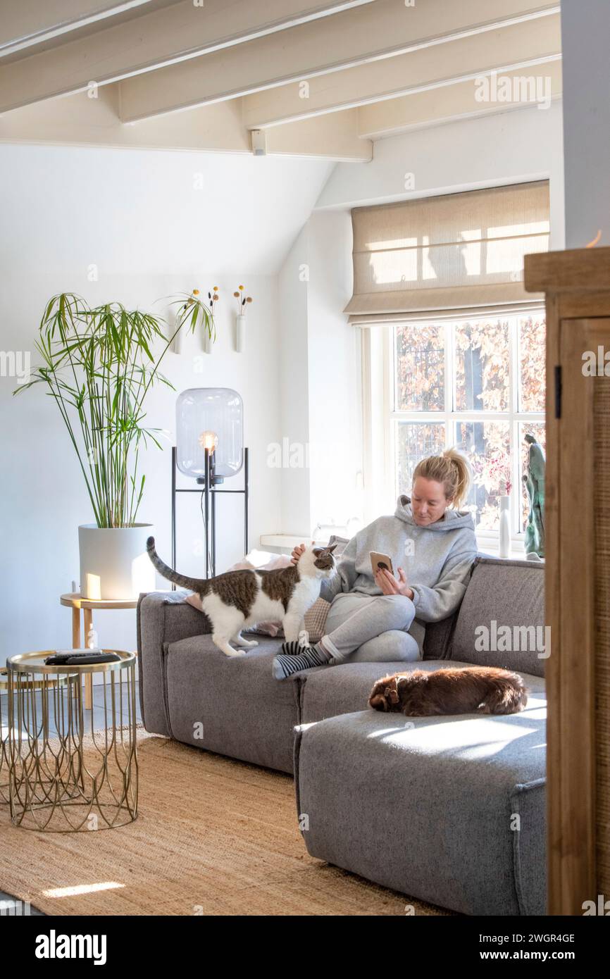 Jeune femme assise sur le canapé dans une maison moderne propre avec son téléphone naviguant avec un feu chaud en arrière-plan et son chat sur ses genoux Banque D'Images