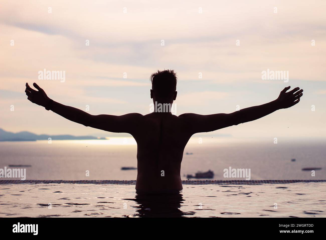 Silhouette d'un jeune homme heureux avec les bras tendus dans la piscine à débordement profitant de la vue au coucher du soleil. Banque D'Images