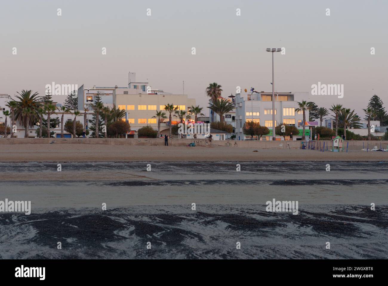 Le coucher du soleil se reflète dans les fenêtres des bâtiments en face d'une plage de sable avec aire de jeux pour enfants clôturée à Essaouira, Maroc, le 6 février 2024 Banque D'Images