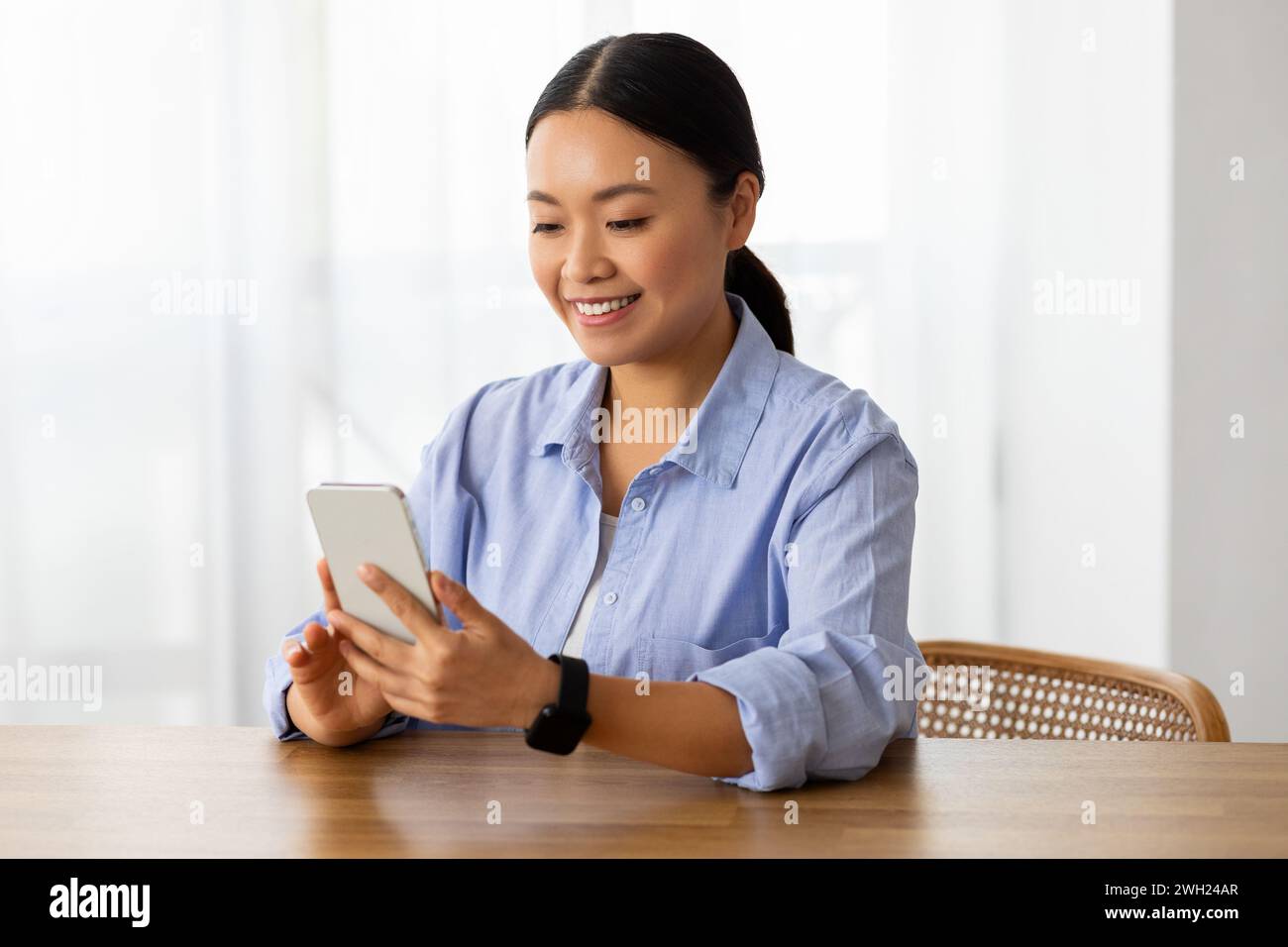 Femme asiatique souriante assise au bureau à la maison, en utilisant le téléphone Banque D'Images