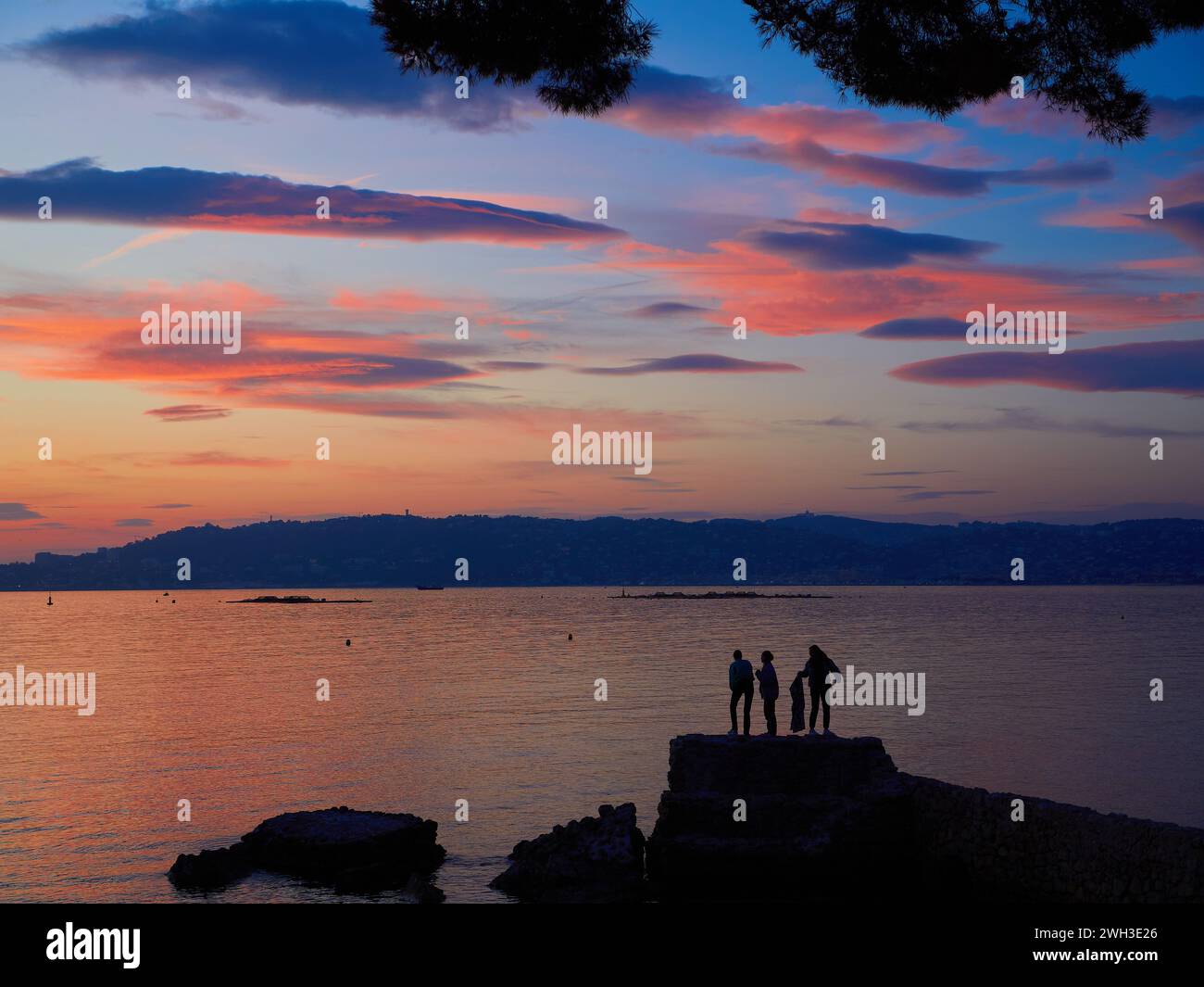 Coucher de soleil nuageux sur la baie de Cannes, France, avec des gens sur les rochers silhouettés contre la mer rose calme. Banque D'Images