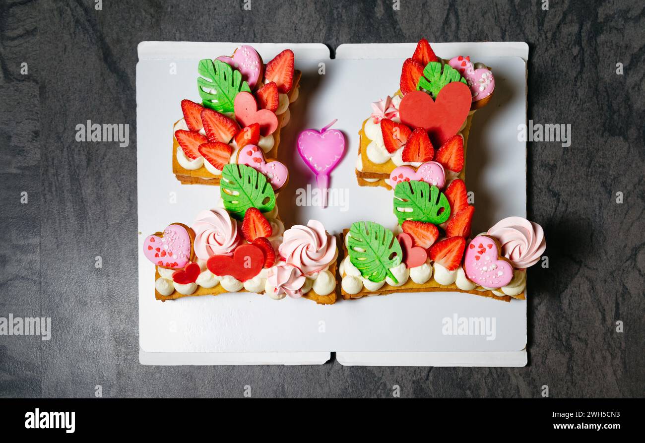 Un gâteau de mille-feuilles de fraise en forme de numéro 11 élaboré, orné de décorations de coeur, de feuilles fondantes vertes et de crème fouettée, présenté dans un w Banque D'Images