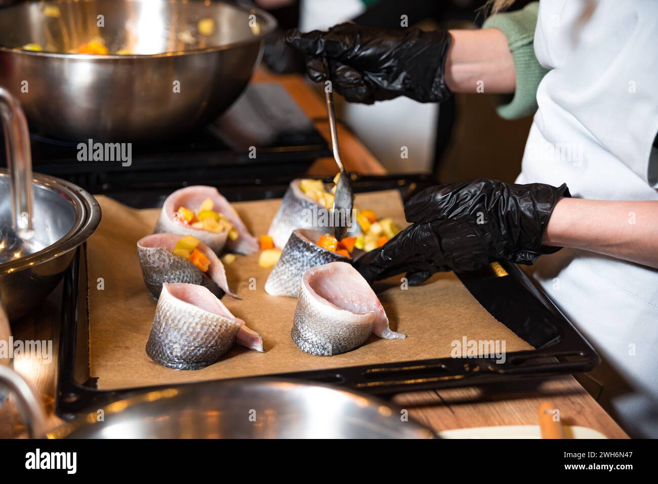 Chef avec des gants noirs farçant soigneusement les filets de poisson avec des légumes coupés en dés pour un repas gastronomique, montrant des techniques culinaires professionnelles. Banque D'Images