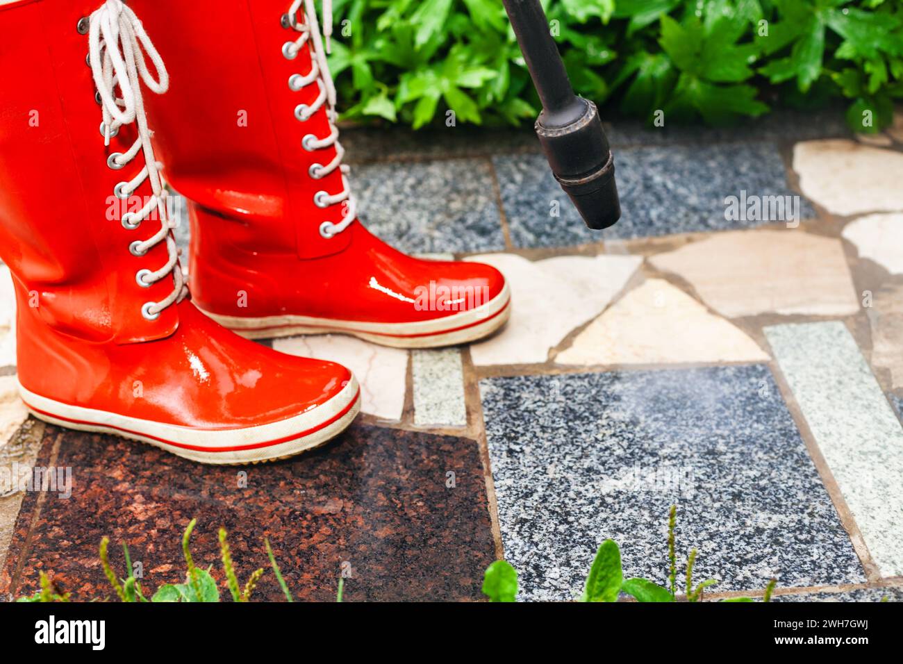 La personne portant des bottes en caoutchouc rouge vif utilise un nettoyeur haute pression pour nettoyer la saleté d'une passerelle de jardin en pierre Banque D'Images