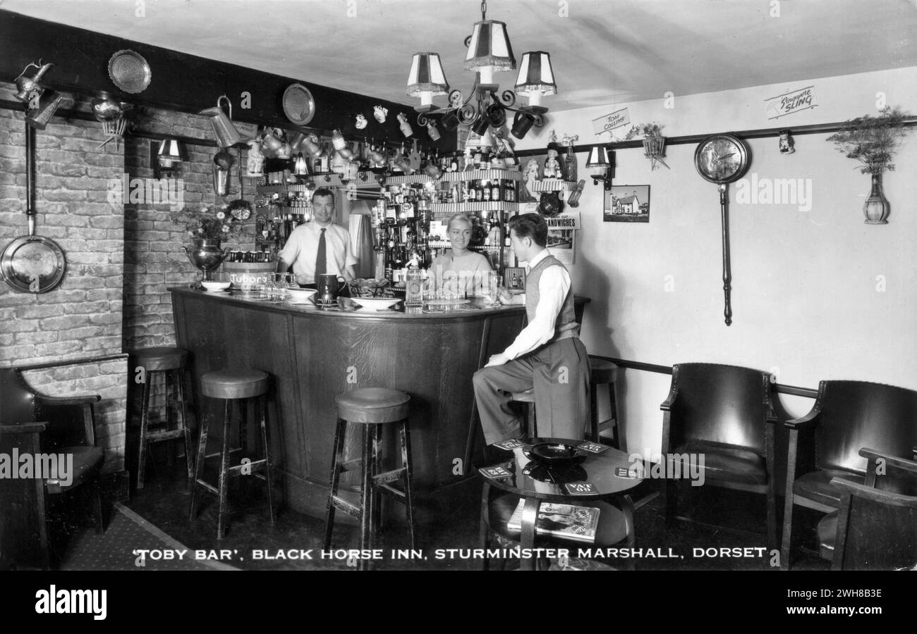 Dorset. c.1960 – Une vraie carte postale photographique intitulée « Toby Bar, Black Horse Inn, Sturminster Marshall, Dorset » représentant le « Toby Bar » dans le Black Horse Inn, une ancienne auberge de coaching située à Sturminster Marshall sur la route A350 entre Poole et Blandford Forum. Le bar est décoré avec une gamme de cruches et de tankards, y compris quelques cruches toby. Les propriétaires, John et Jean Sibley sont debout derrière le bar. Banque D'Images