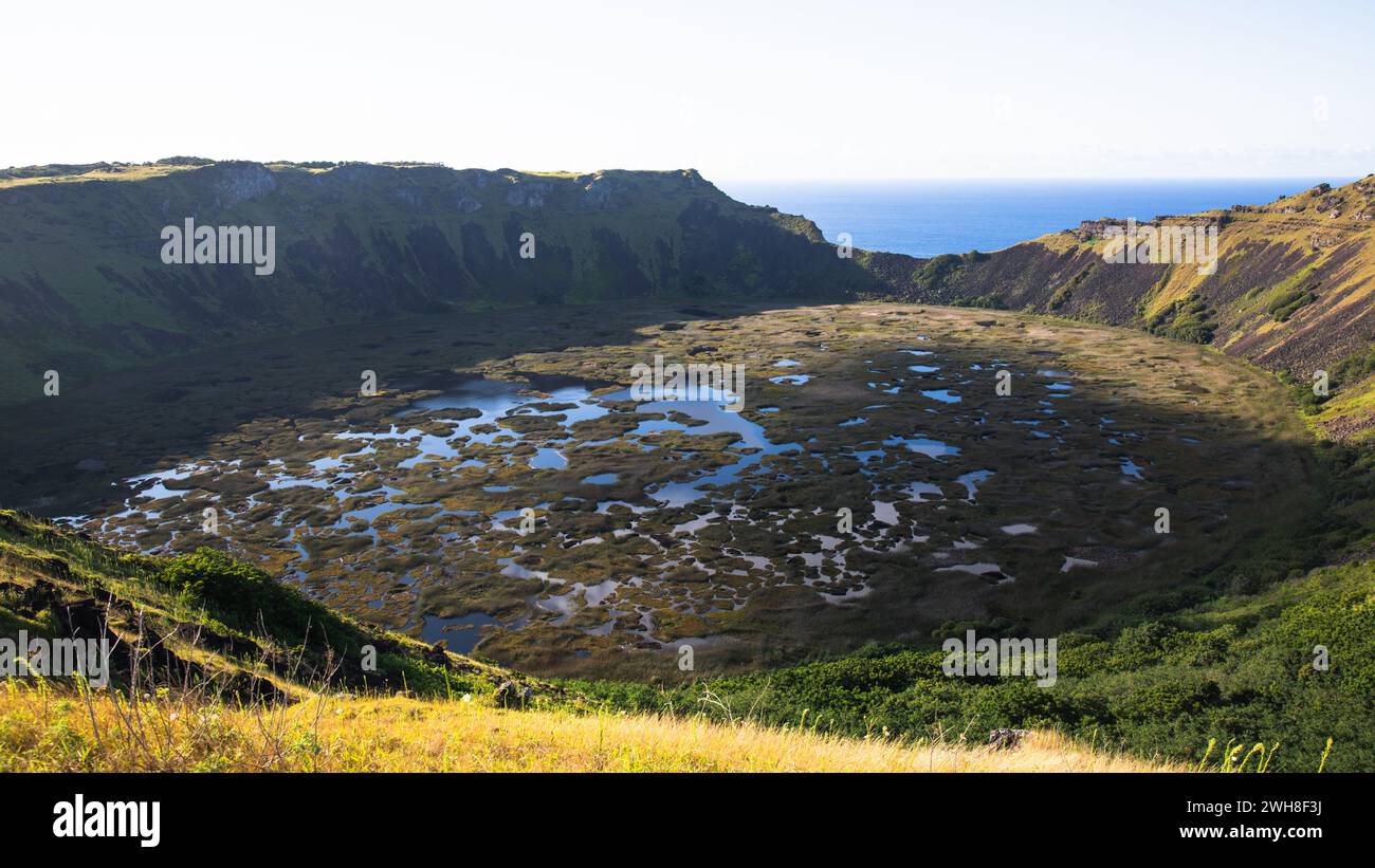 Paysage marécageux avec des trous d'eau remplis au pied du volcan Rano Kau et l'océan Pacifique en arrière-plan. Banque D'Images